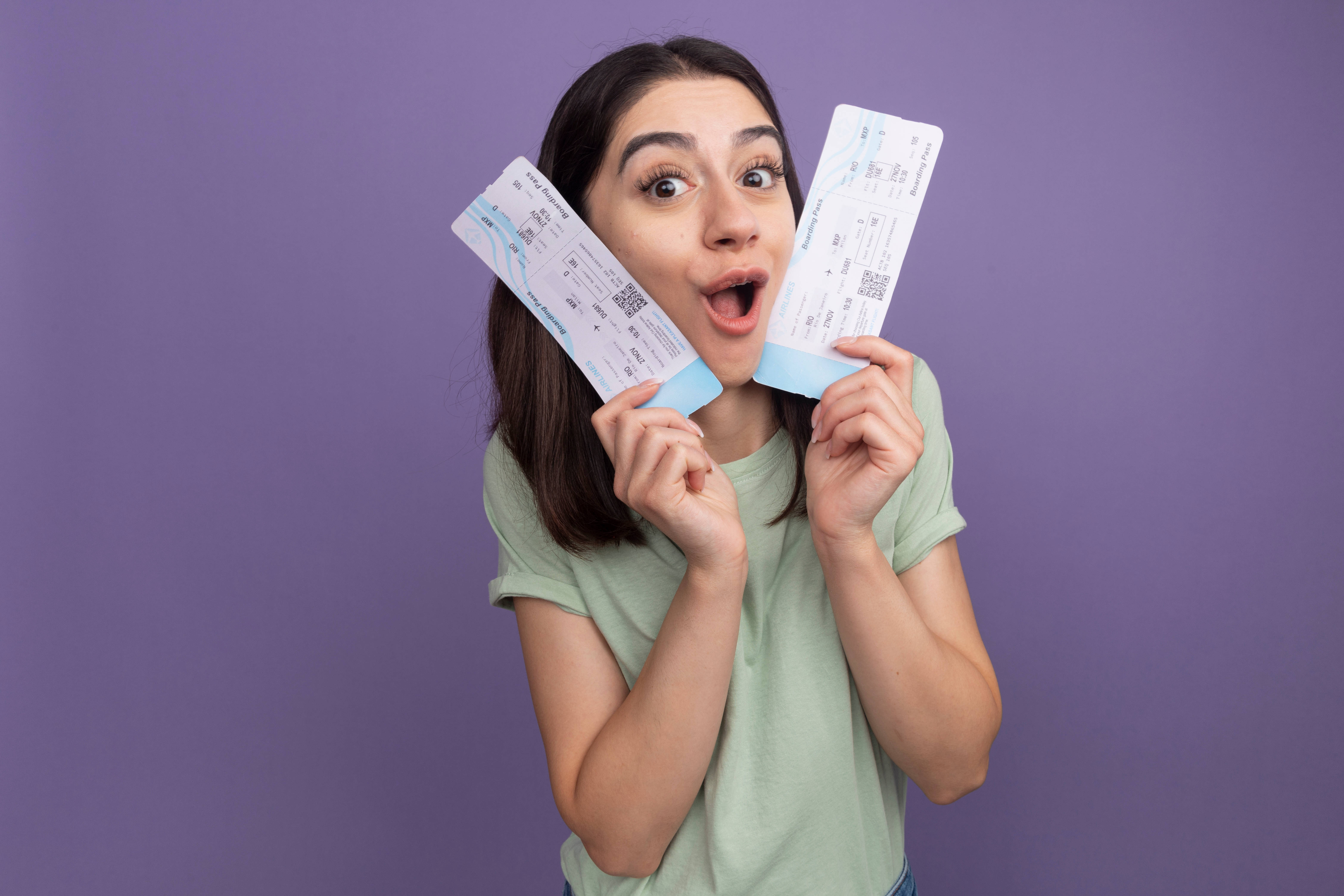 Impressed young pretty caucasian girl holding airplane tickets touching face with them