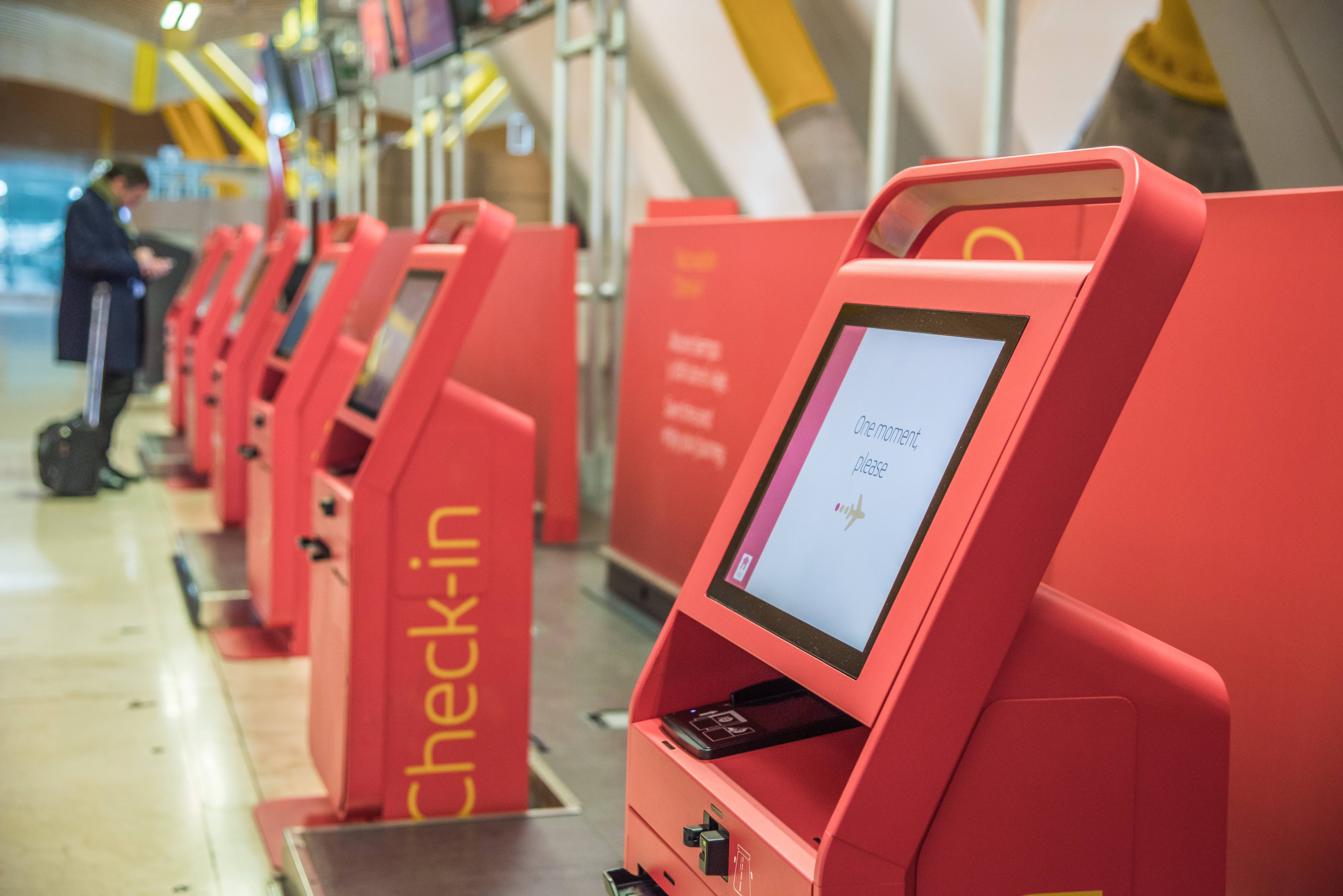 Red self-service check-in kiosks at an airport terminal, with passengers using the machines