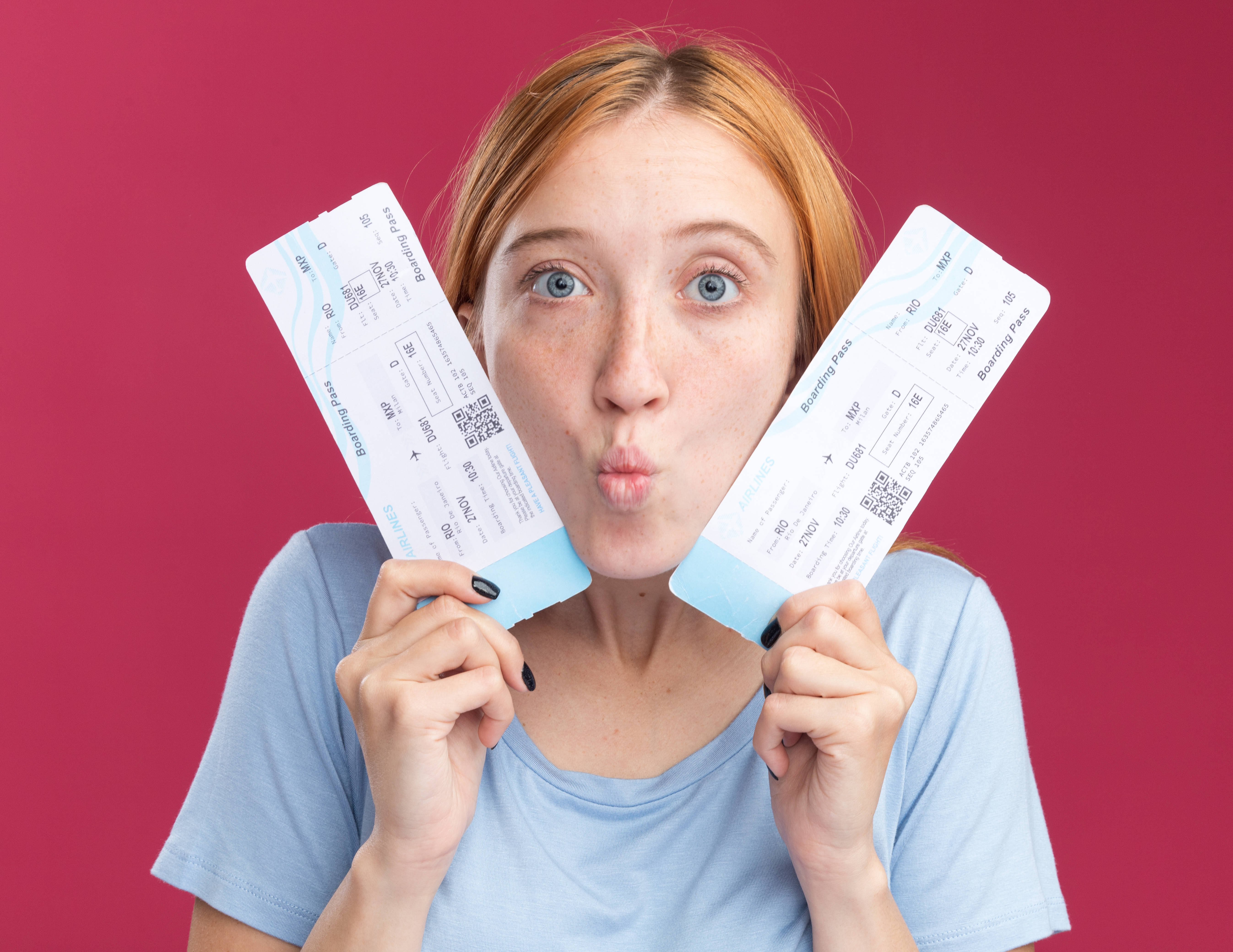 Surprised young redhead ginger girl with freckles holding air tickets close to face on pink