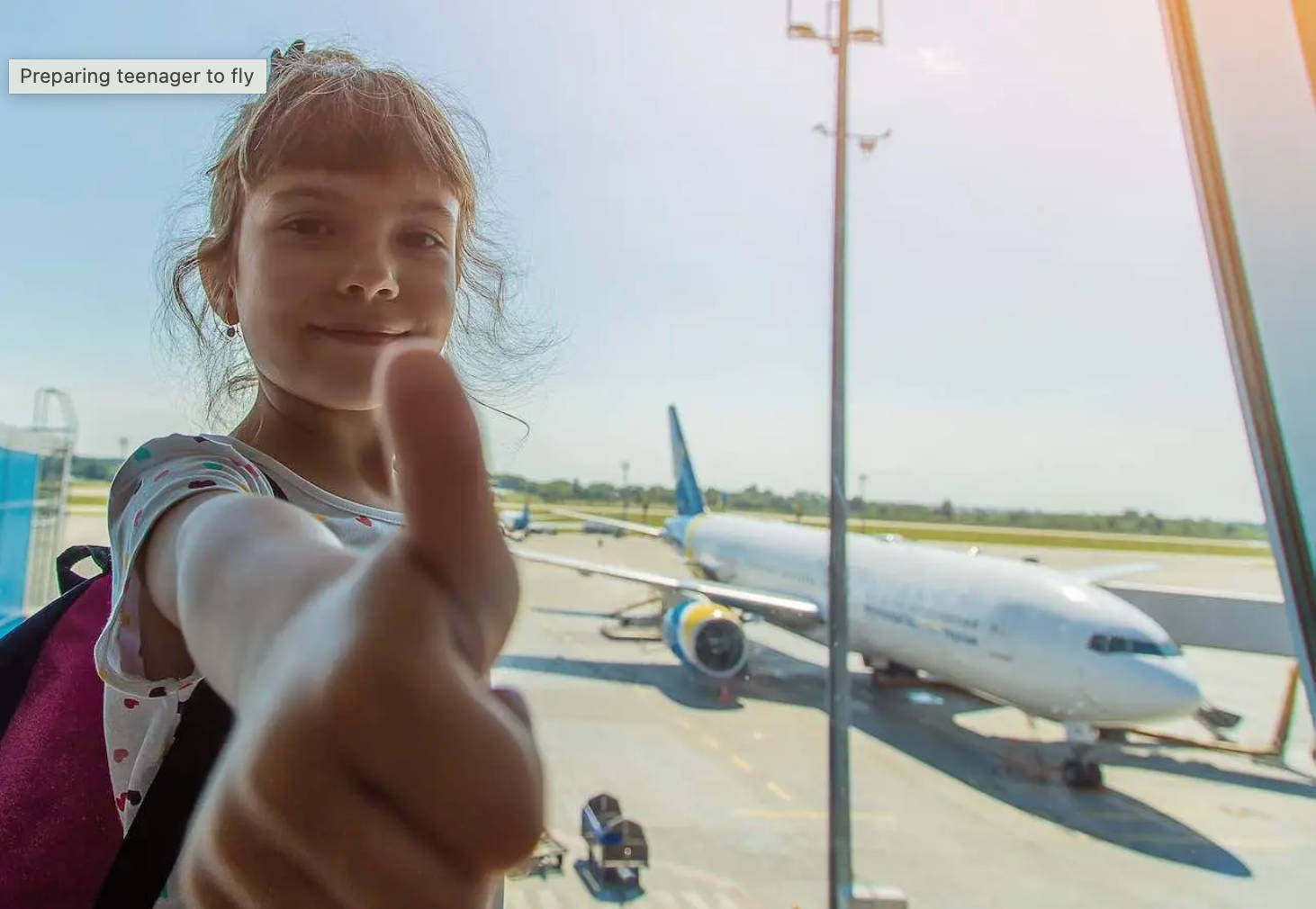 young girl stands in terminal with thumb up, plane in background