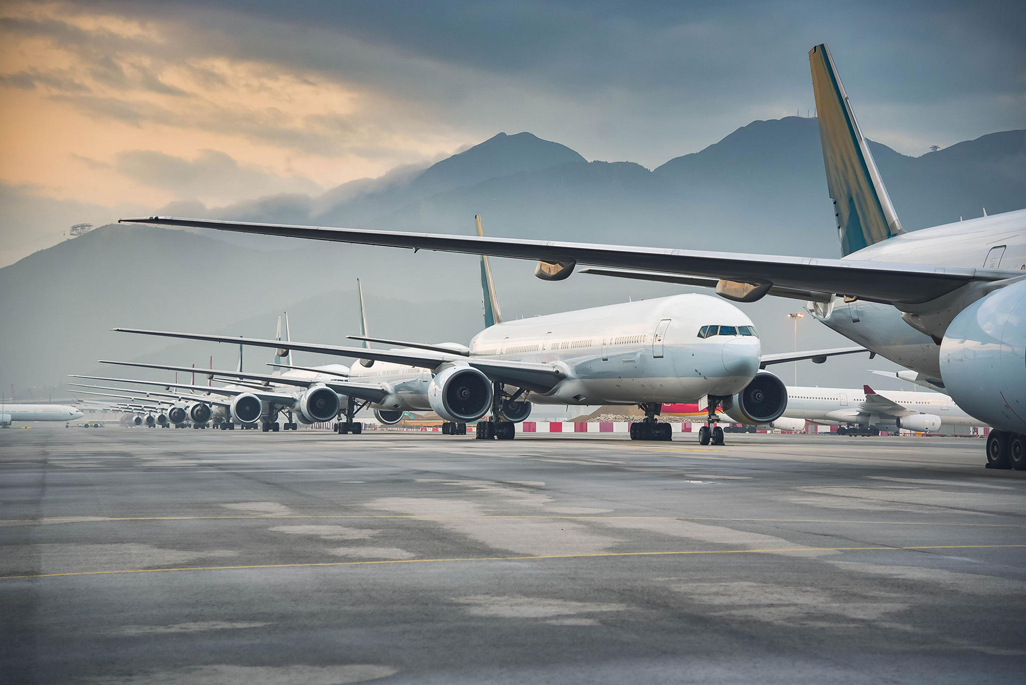 A row of parked commercial airplanes on a tarmac with mountains in the background under a cloudy sky. The planes' large wingspans are prominent, creating a serene, overcast scene